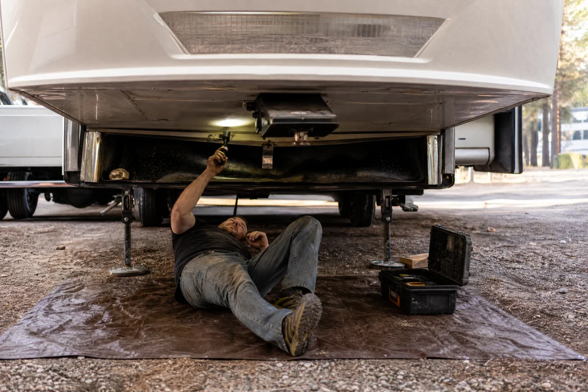 Technician lying under RV with flashlight inspecting undercarriage for seasonal maintenance and pest prevention.
